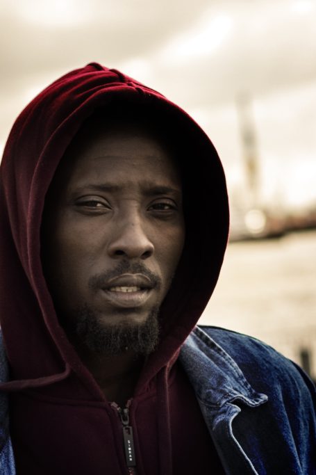 Mann mit Kapuzenpulli und blauer Jacke, ernst blickend, unscharfer Hintergrund.